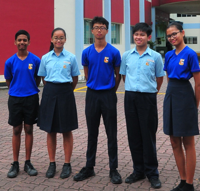 Five school students in blue uniforms stand together outdoors in front of a red and white building.